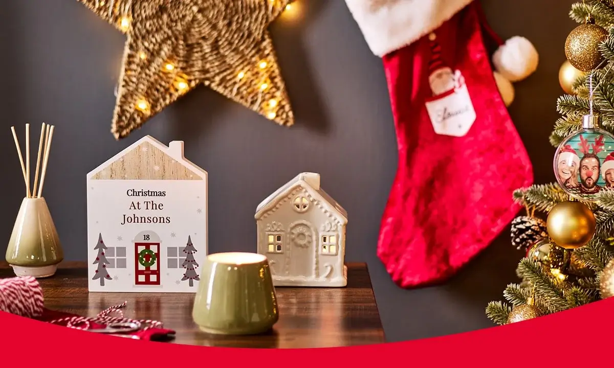 Festive Christmas scene with a decorated tree, red stocking, personalized house sign, ceramic house, lit star ornament, reed diffuser, candle holder, and wrapped gifts Festive Christmas scene with a decorated tree, red stocking, personalized house sign, ceramic house, lit star ornament, reed diffuser, candle holder, and wrapped gifts