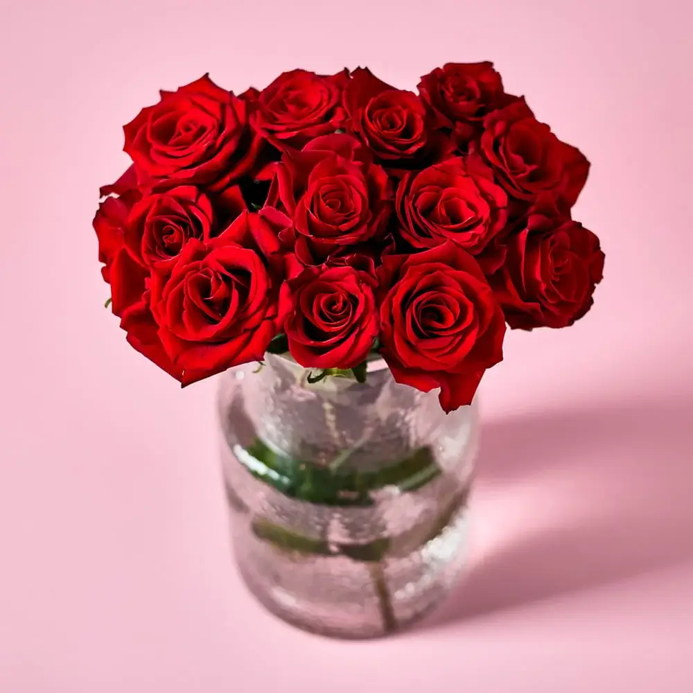 A bright bouquet of yellow roses, white blooms, and greenery arranged in a ribbed white vase, with additional flowers displayed in an open box behind it on a soft pink background.