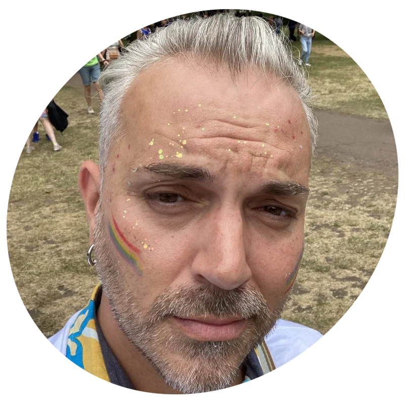 A close‑up selfie of a person outdoors at an event, with short grey hair and a beard, wearing face paint including a small rainbow stripe on the cheek and gold glitter on the face, with people and grass visible in the background.