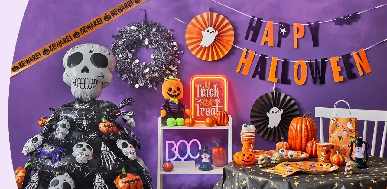 Halloween-themed party setup with a purple backdrop, black netting, paper bats, and garlands featuring pumpkins, skulls, and cat faces, centered around a large black cat face decoration with BOO and SPOOKY text, and a table set with themed plates, cups, candy, and spooky decorations.