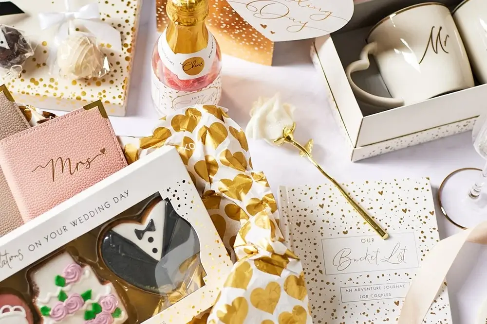 'A wedding-themed flat lay featuring various items including a pink notebook with the word 'Mrs.' in gold script, a bottle of champagne labeled 'Cheers', heart-shaped cookies, a white mug with an 'M' on it, and a journal titled 'Our Bucket List: An Adventure Journal for Couples'. The items are surrounded by gold and white wrapping paper with heart patterns.' 'A wedding-themed flat lay featuring various items including a pink notebook with the word 'Mrs.' in gold script, a bottle of champagne labeled 'Cheers', heart-shaped cookies, a white mug with an 'M' on it, and a journal titled 'Our Bucket List: An Adventure Journal for Couples'. The items are surrounded by gold and white wrapping paper with heart patterns.'