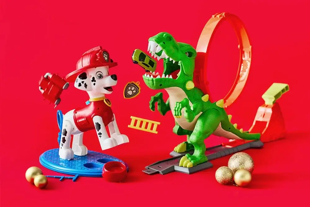 Colorful toy setup featuring a Dalmatian firefighter figure from Paw Patrol and a green dinosaur with an open mouth holding a small car, displayed against a bright red background with gold ornaments and play accessories.