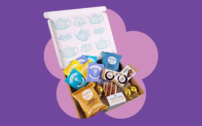 A themed treat box filled with biscuits, cakes, hot drink sachets, and chocolates, shown with the box lid open against a purple background with a pink flower shape.
