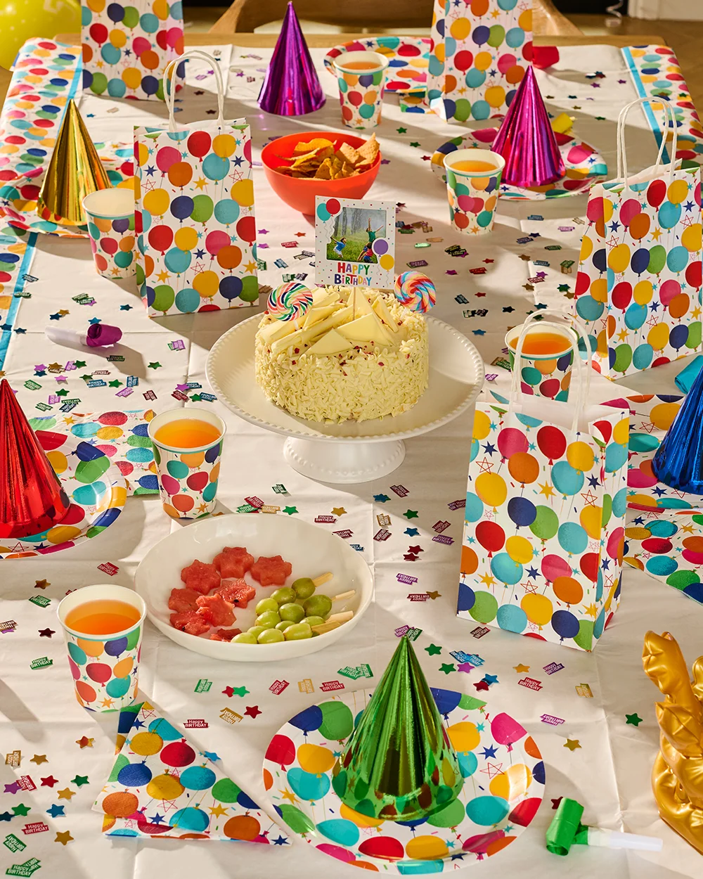 A colourful birthday party table decorated with balloon-print partyware, including matching plates, cups, napkins and gift bags. The display features metallic party hats in bright colours, scattered confetti, party blowers, snacks, drinks, and a birthday cake topped with a ‘Happy Birthday’ decoration, creating a vibrant celebration setup. A colourful birthday party table decorated with balloon-print partyware, including matching plates, cups, napkins and gift bags. The display features metallic party hats in bright colours, scattered confetti, party blowers, snacks, drinks, and a birthday cake topped with a ‘Happy Birthday’ decoration, creating a vibrant celebration setup.