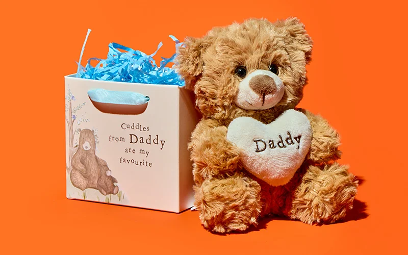 A soft brown teddy bear holding a cream heart‑shaped cushion embroidered with ‘Daddy,’ displayed beside a keepsake gift box printed with the message ‘Cuddles from Daddy are my favourite,’ against a bright orange background with blue shredded paper inside the box. A soft brown teddy bear holding a cream heart‑shaped cushion embroidered with ‘Daddy,’ displayed beside a keepsake gift box printed with the message ‘Cuddles from Daddy are my favourite,’ against a bright orange background with blue shredded paper inside the box.