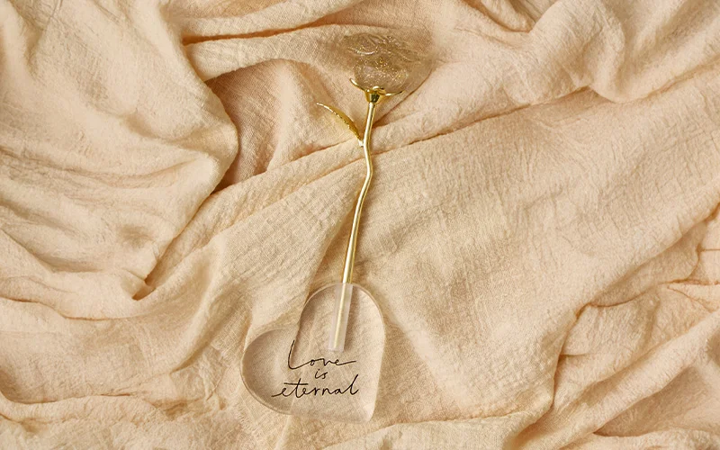 A decorative glass rose with a gold stem and a clear heart base engraved with the words ‘Love is eternal,’ placed on soft beige fabric. A decorative glass rose with a gold stem and a clear heart base engraved with the words ‘Love is eternal,’ placed on soft beige fabric.