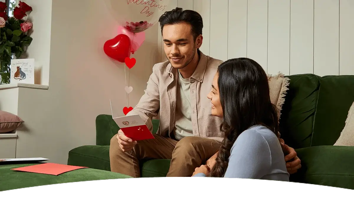 Couple sitting together in a cosy living room decorated with Valentine’s balloons, as one person holds an open card.