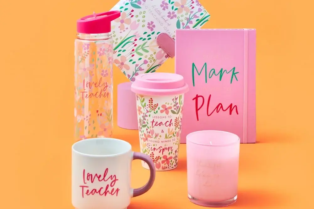 'The image shows a collection of colorful teacher-themed items against an orange background. The items include a clear water bottle with pink accents and the text 'Lovely Teacher', a white mug with the same text, a pink travel cup with floral designs and the text 'Lessons to teach & young minds to inspire', a pink candle with faint gold text, another candle in the background, a floral-patterned notebook, and a pink planner with the words 'Mark Plan' on its cover.' 'The image shows a collection of colorful teacher-themed items against an orange background. The items include a clear water bottle with pink accents and the text 'Lovely Teacher', a white mug with the same text, a pink travel cup with floral designs and the text 'Lessons to teach & young minds to inspire', a pink candle with faint gold text, another candle in the background, a floral-patterned notebook, and a pink planner with the words 'Mark Plan' on its cover.'