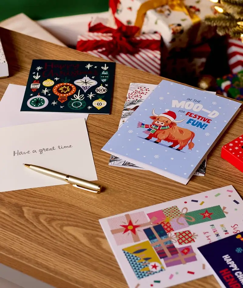 Selection of Christmas cards and festive stationery displayed on a wooden table with wrapped presents in the background. Selection of Christmas cards and festive stationery displayed on a wooden table with wrapped presents in the background.