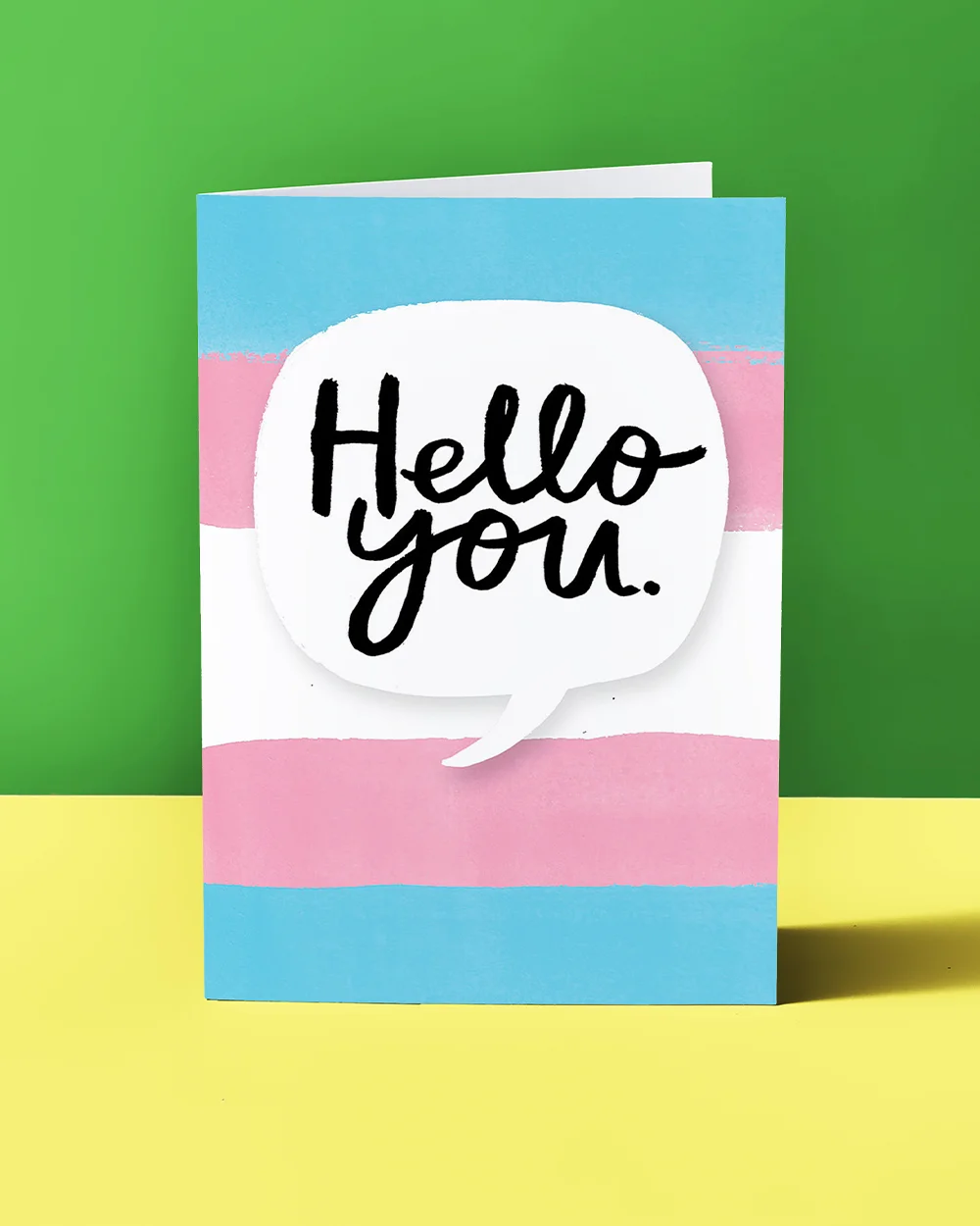 A greeting card with horizontal blue, pink and white stripes inspired by the trans pride flag, featuring a white speech bubble with bold black script text reading ‘Hello you,’ displayed against a green and yellow background.