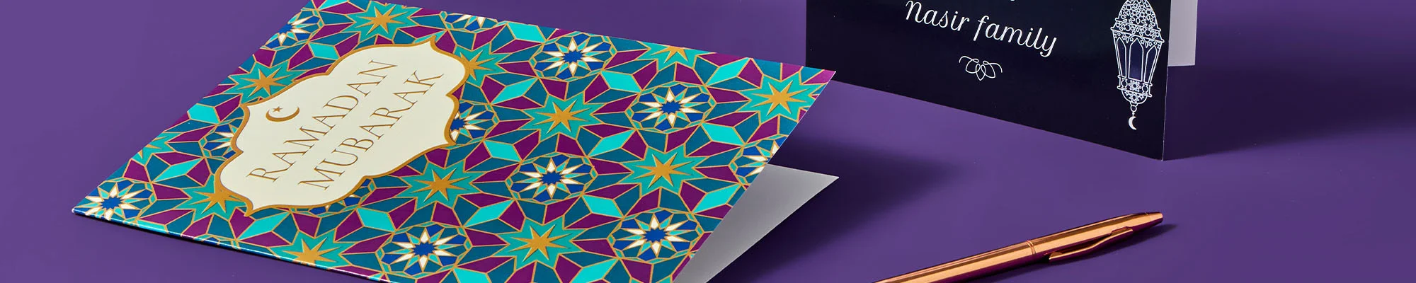 A set of Ramadan and Eid greeting cards displayed on a purple background, including a colourful geometric patterned card and a dark card with lantern artwork, with a pen placed beside them.