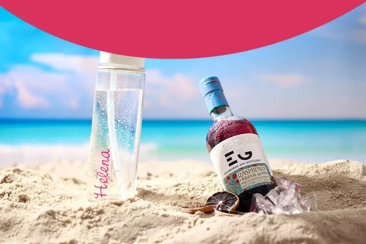 A clear water bottle with the name Helena written in pink is placed on sandy beach next to a bottle of Edinburgh Gin Distillery Raspberry Liqueur. The background shows a serene ocean view with blue skies and soft clouds. There are also some ice cubes and dried citrus slices near the gin bottle. A clear water bottle with the name Helena written in pink is placed on sandy beach next to a bottle of Edinburgh Gin Distillery Raspberry Liqueur. The background shows a serene ocean view with blue skies and soft clouds. There are also some ice cubes and dried citrus slices near the gin bottle.