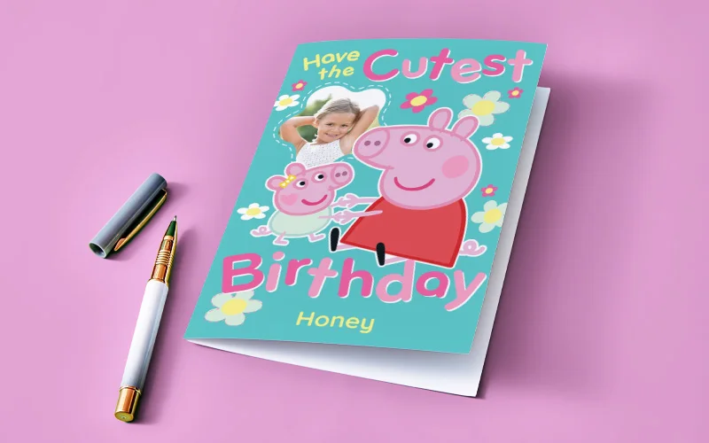 A personalised Peppa Pig birthday card with a turquoise background and flower illustrations, featuring Peppa Pig characters, a photo window, and the text ‘Have the Cutest Birthday,’ with the name ‘Honey’ printed at the bottom. A pen is placed beside the card on a pink background.