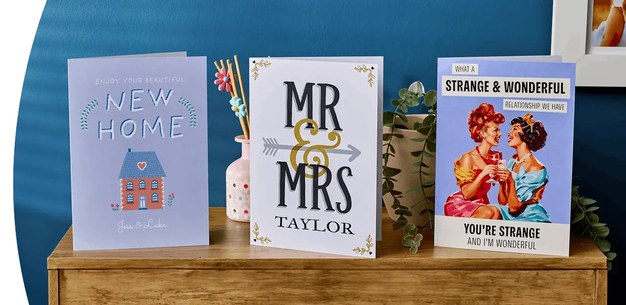 Three greeting cards displayed on a wooden surface, including a new home card, a personalised Mr and Mrs wedding card, and a humorous vintage-style relationship card against a blue background.