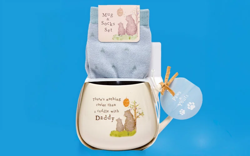A ceramic mug and socks gift set displayed on a blue background, featuring a cream mug printed with an illustration of a bear and cub and the text ‘There’s nothing cosier than a cuddle with Daddy,’ with a matching pair of light blue socks and a small tag reading ‘Days with you,’ arranged together. A ceramic mug and socks gift set displayed on a blue background, featuring a cream mug printed with an illustration of a bear and cub and the text ‘There’s nothing cosier than a cuddle with Daddy,’ with a matching pair of light blue socks and a small tag reading ‘Days with you,’ arranged together.