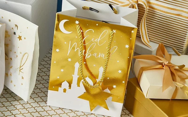 Gold and white Eid gift bag decorated with a crescent moon, stars, and mosque silhouettes, with gold rope handles and a star‑shaped gift tag.