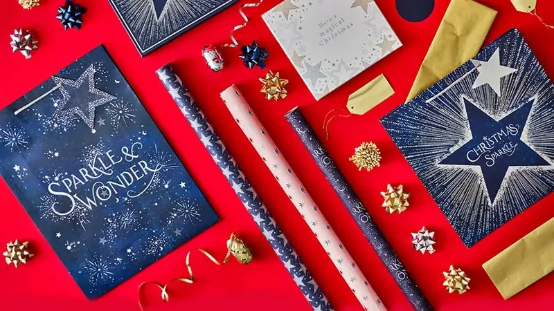 Christmas gift wrapping set displayed on a red background, featuring navy and gold gift bags with star designs and text Sparkle and Wonder and Christmas Sparkle, coordinating wrapping paper rolls in blue and pink patterns, gold envelopes, decorative bows, and curling ribbons.