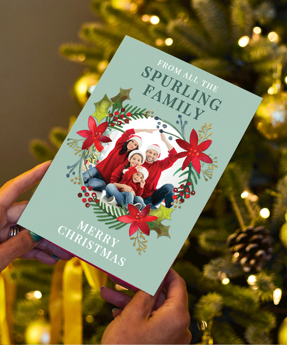 Christmas card held in front of a decorated tree, featuring a family photo framed by a festive wreath with red poinsettias and holly, with the text From all the Spurling Family at the top and Merry Christmas at the bottom.