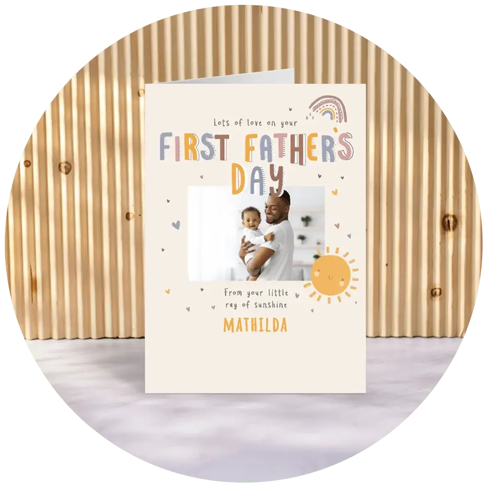 A personalised first Father’s Day card with a cream background, featuring illustrated suns, hearts and a rainbow, the text ‘Lots of love on your First Father’s Day,’ and a square photo window with a personal name printed below, displayed against a wooden slatted backdrop. A personalised first Father’s Day card with a cream background, featuring illustrated suns, hearts and a rainbow, the text ‘Lots of love on your First Father’s Day,’ and a square photo window with a personal name printed below, displayed against a wooden slatted backdrop.