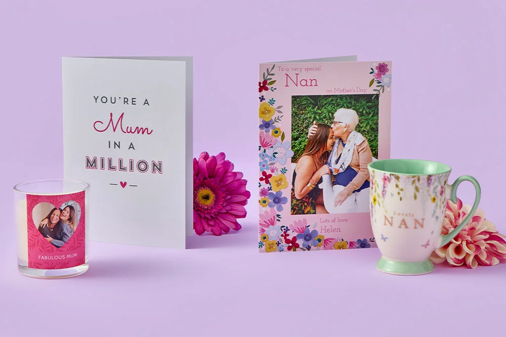 Close‑up display of Mother’s Day items on a purple background, including two greeting cards, a floral mug labelled ‘Nan,’ a personalised candle, and bright pink flowers.