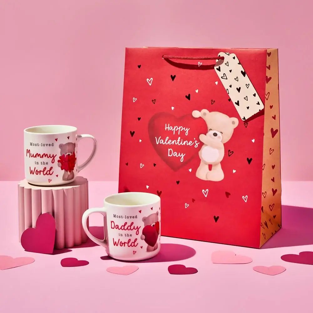Red Valentine’s Day gift bag with a teddy bear design and heart patterns, accompanied by two white mugs with red text reading Most-loved Mummy in the World and Most-loved Daddy in the World, surrounded by scattered paper hearts on a pink background.