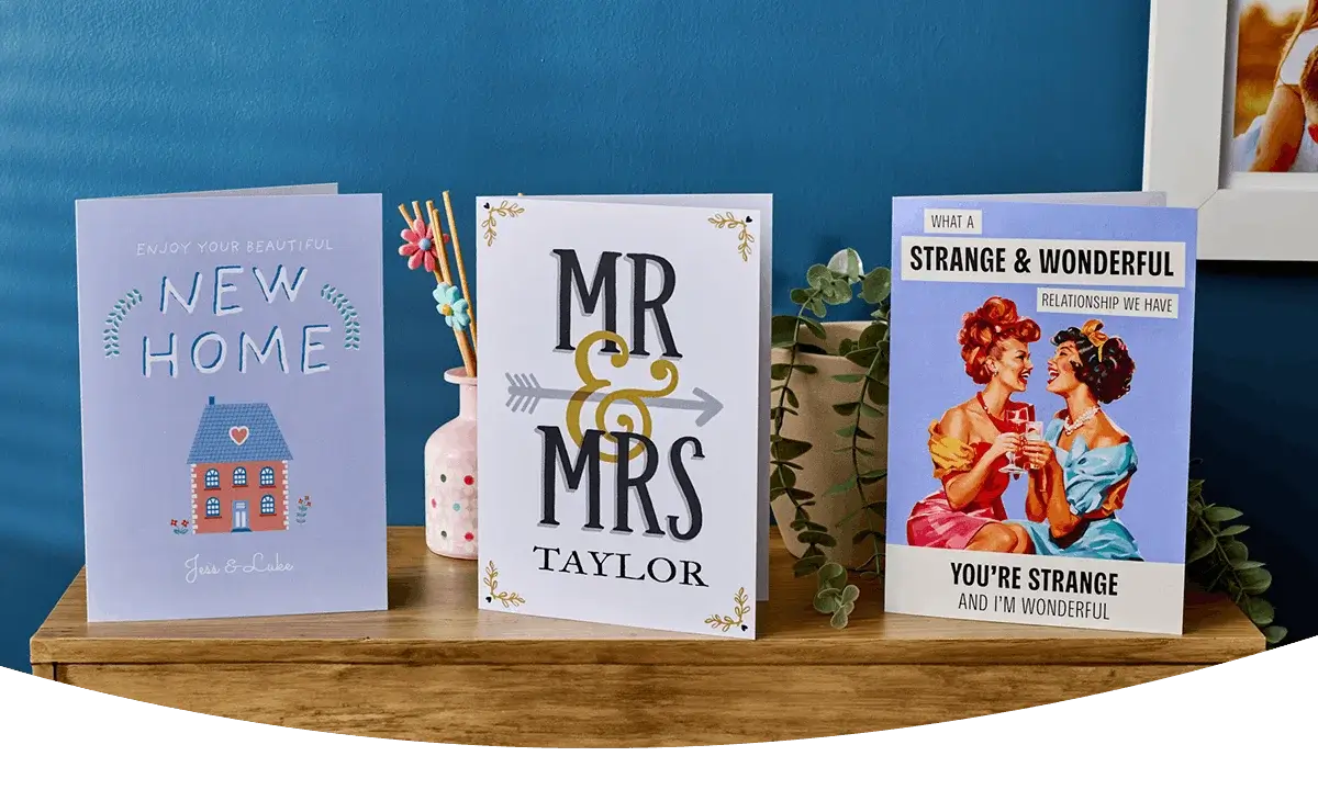 Three greeting cards displayed on a wooden surface, including a new home card, a personalised Mr and Mrs wedding card, and a humorous vintage-style relationship card against a blue background.