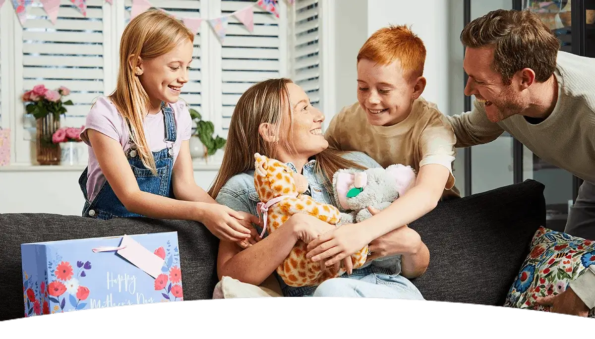 A family gathered on a sofa celebrating Mother’s Day, with children giving gifts and plush toys while decorations and flowers brighten the room.