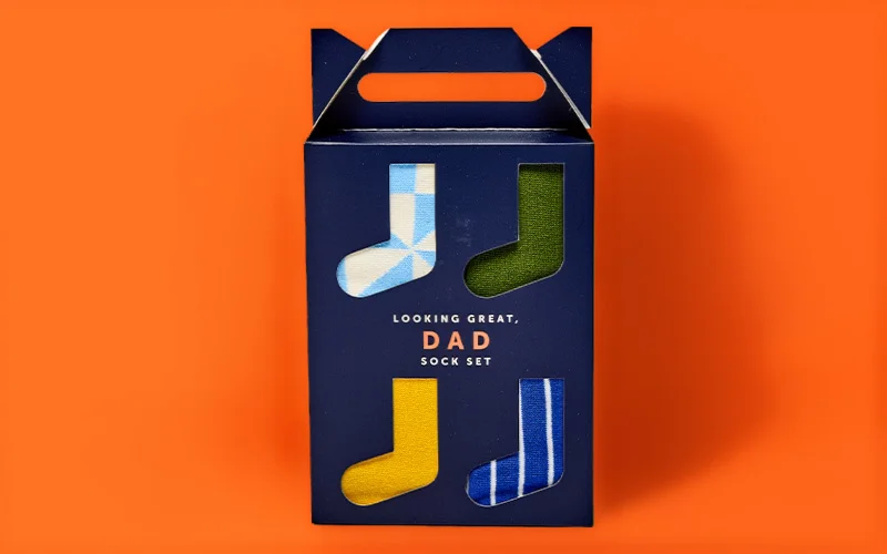 A boxed ‘Dad’ sock gift set in navy packaging with cut‑out windows showing four pairs of socks in different colours and patterns, with the text ‘Looking Great, Dad Sock Set’ on the front, displayed against a bright orange background. A boxed ‘Dad’ sock gift set in navy packaging with cut‑out windows showing four pairs of socks in different colours and patterns, with the text ‘Looking Great, Dad Sock Set’ on the front, displayed against a bright orange background.