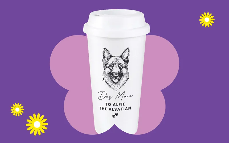 A white travel cup featuring a detailed illustration of a dog and the text ‘Dog Mum – To Alfie the Alsatian,’ shown against a purple background with a pink flower shape and yellow daisies.