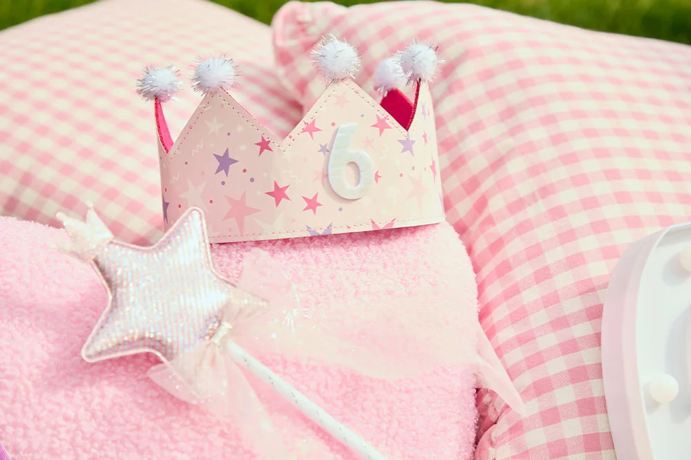 A pastel pink princess crown with star illustrations and a white number 6 on the front, resting on a soft pink blanket beside a shimmering star-shaped wand, with pink gingham cushions in the background. A pastel pink princess crown with star illustrations and a white number 6 on the front, resting on a soft pink blanket beside a shimmering star-shaped wand, with pink gingham cushions in the background.