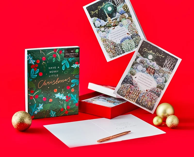 Set of Christmas cards on a bright red background, including a box labeled Have a Merry Little Christmas with festive foliage and berries, two propped-up cards showing snowy winter scenes with a church and a house, gold ornaments, and an open card with an envelope and pen.