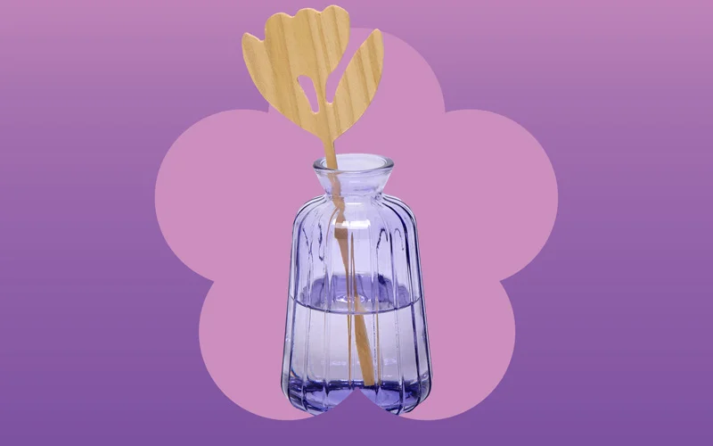 A clear ribbed glass vase filled partially with liquid, holding a wooden flower-shaped diffuser stick, displayed against a purple and pink gradient background with a flower silhouette.