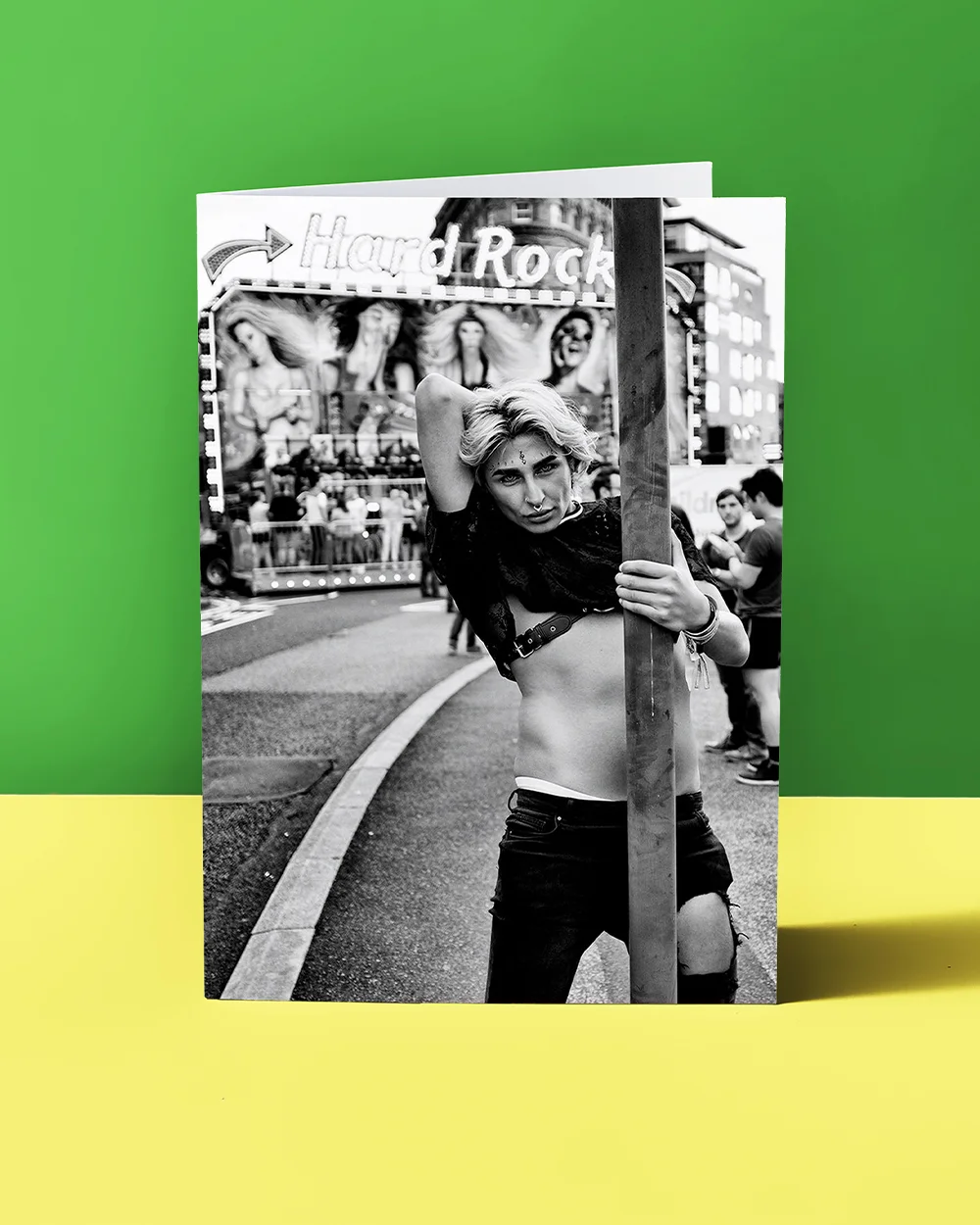 A black‑and‑white photographic greeting card showing a person posing while holding a vertical pole in an outdoor fairground setting, with a ‘Hard Rock’ sign and amusement ride visible in the background.