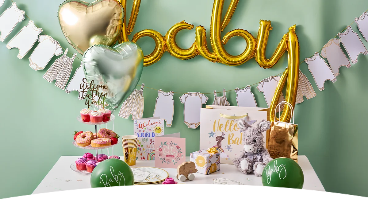 A baby shower party table decorated with gold letter balloons spelling ‘Baby’, baby-clothes bunting and heart-shaped foil balloons against a sage green backdrop. The display features pink cupcakes, doughnuts, baby shower cards, a ‘Hello Baby’ gift bag, a plush donkey toy, green balloons printed with ‘Baby,’ and gold number-shaped balloons, creating a coordinated baby shower celebration setup. A baby shower party table decorated with gold letter balloons spelling ‘Baby’, baby-clothes bunting and heart-shaped foil balloons against a sage green backdrop. The display features pink cupcakes, doughnuts, baby shower cards, a ‘Hello Baby’ gift bag, a plush donkey toy, green balloons printed with ‘Baby,’ and gold number-shaped balloons, creating a coordinated baby shower celebration setup.