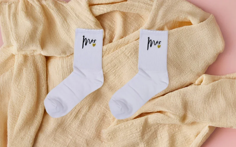A pair of white socks with ‘Mrs’ printed in black script and a small gold heart detail, laid flat on soft beige fabric. A pair of white socks with ‘Mrs’ printed in black script and a small gold heart detail, laid flat on soft beige fabric.