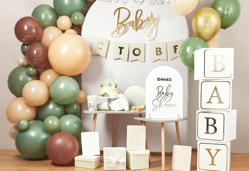 A baby shower celebration display featuring gold ‘Baby’ balloons, baby-clothes bunting, pastel green and gold balloons, baby-themed greeting cards, wrapped gifts, a plush donkey toy and coordinating tableware arranged on a decorated party table.