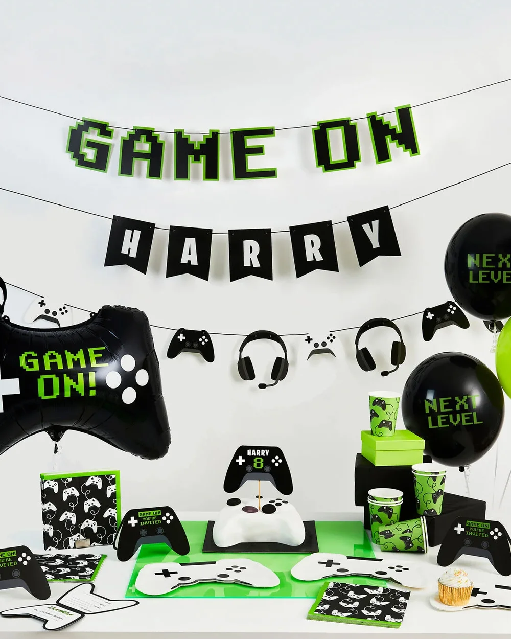 A gaming-themed birthday party display featuring black, white and neon green decorations, including a personalised ‘GAME ON’ banner with the name ‘Harry,’ video game controller bunting, ‘Next Level’ balloons, themed cups and napkins, and a controller-shaped birthday cake. The table is styled with gaming partyware, invitations and controller decorations, with a large game-controller foil balloon displayed to one side.