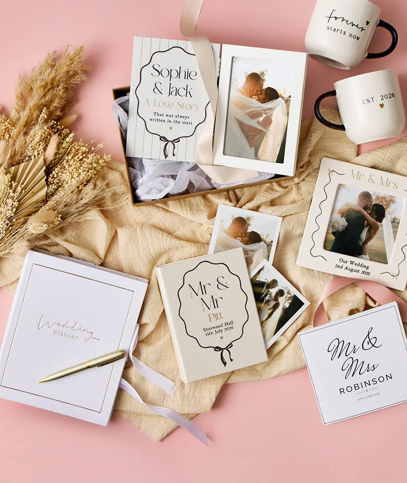 Wedding-themed stationery and personalised gifts including photo cards, mugs, keepsake boxes, and a wedding planner arranged on a soft pink background