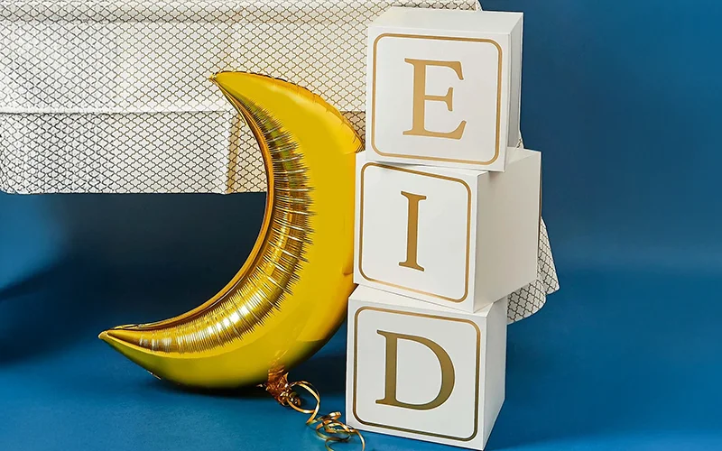 White stacking blocks with gold letters spelling ‘EID’ displayed beside a gold crescent‑moon balloon against a blue background.