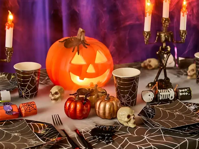 Halloween party table featuring a glowing pumpkin lantern, spiderweb tableware, decorative pumpkins, skull ornaments and spooky candle holders.