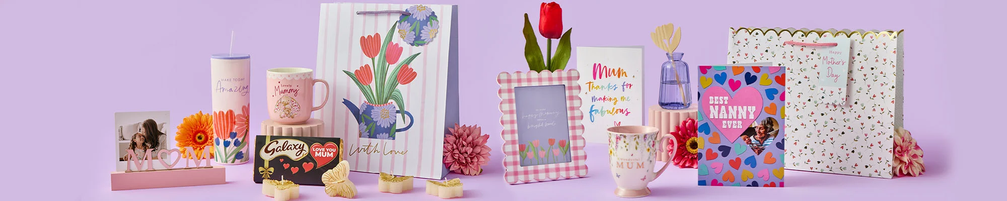 A wide display of Mother’s Day gifts arranged on a pastel purple background, including floral gift bags, greeting cards, a framed photo holder, mugs, chocolates, a tumbler, artificial flowers, and small decorative items.
