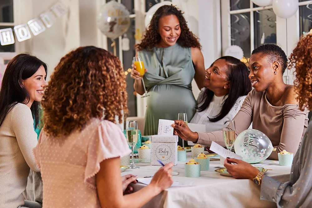 A baby shower celebration with guests seated around a decorated table filled with cupcakes, drinks and baby shower games. The display includes white balloons, baby-themed decorations, a ‘Hello Baby’ balloon and game cards, creating a relaxed baby shower party setting. A baby shower celebration with guests seated around a decorated table filled with cupcakes, drinks and baby shower games. The display includes white balloons, baby-themed decorations, a ‘Hello Baby’ balloon and game cards, creating a relaxed baby shower party setting.