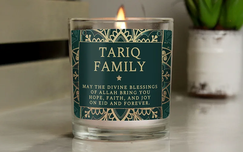 A lit candle in a glass jar featuring a green and gold patterned label personalised with ‘Tariq Family’ and a printed Eid blessing.