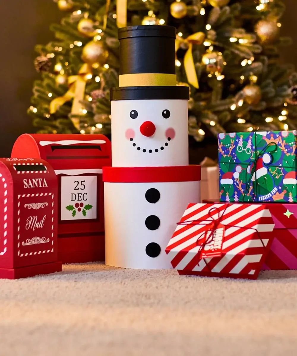 Christmas gift boxes and a snowman-shaped container placed in front of a decorated Christmas tree with golden ornaments and lights.