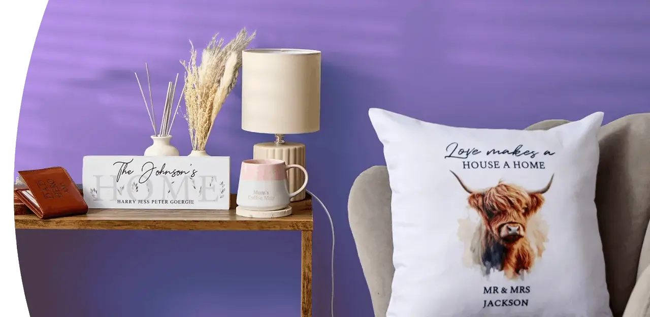 A cosy home display featuring personalised wall art, a wooden clock, a table lamp, framed prints, decorative accessories and a cushion with a highland cow design arranged against a dark blue wall.