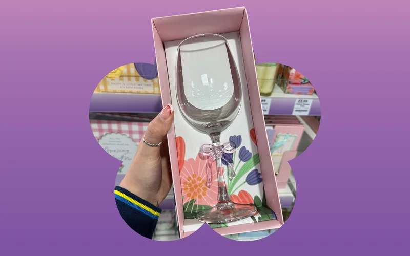 A clear wine glass presented in a pink gift box with colourful floral illustrations, held in someone’s hand, displayed against a purple and pink gradient background with a flower silhouette.
