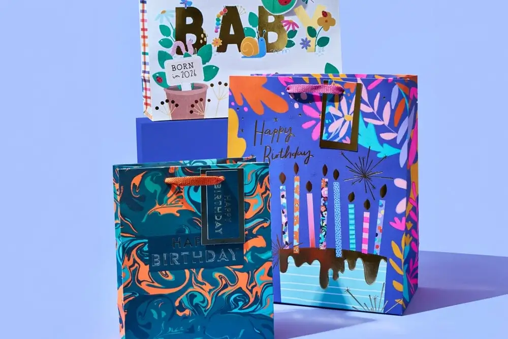'The image shows three colorful gift bags with different designs. The first bag has a blue and orange marbled pattern with the text 'HAPPY BIRTHDAY' embossed on it. The second bag features a vibrant design with candles, flowers, and the text 'Happy Birthday'. The third bag in the background has a baby-themed design with the text 'BABY' and 'BORN IN 2024' 'The image shows three colorful gift bags with different designs. The first bag has a blue and orange marbled pattern with the text 'HAPPY BIRTHDAY' embossed on it. The second bag features a vibrant design with candles, flowers, and the text 'Happy Birthday'. The third bag in the background has a baby-themed design with the text 'BABY' and 'BORN IN 2024'