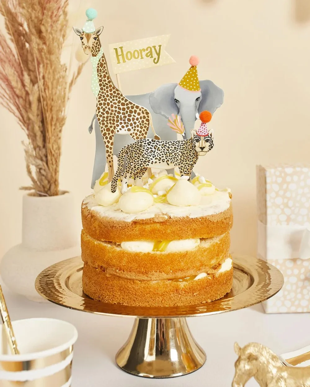 A safari-themed celebration cake displayed on a gold cake stand, decorated with animal toppers including a giraffe, elephant and leopard wearing party hats. A small flag reading ‘Hooray’ sits among the decorations, while cream frosting, cupcakes, gift boxes and safari-themed party accessories surround the cake against a neutral beige backdrop.