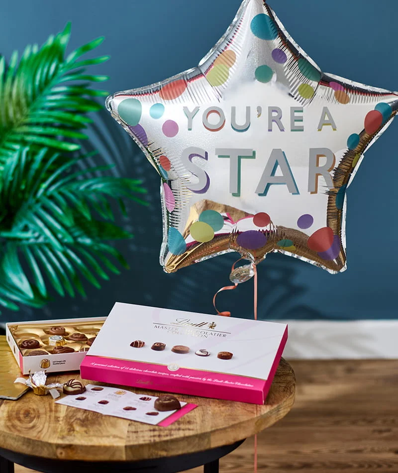 Star-shaped balloon with You’re a Star message alongside a box of chocolates on a wooden table against a blue background