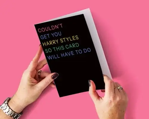 Trending greeting card with colourful text reading Couldn't Get You Harry Styles So This Card Will Have To Do held against a pink background.